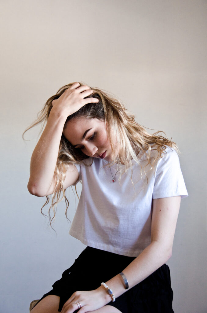 Darian Rojas a 21-year-old from San Francisco California poses for a portrait against a neutral backdrop. She wears a white crop top and a black skirt accessorized with a delicate necklace and a beaded bracelet. Her hand gracefully touches her head and her wavy blonde hair flows freely.
